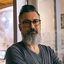 Man with dark hair and beard wearing glasses and a gray t-shirt, sitting in a room with a bright window behind.